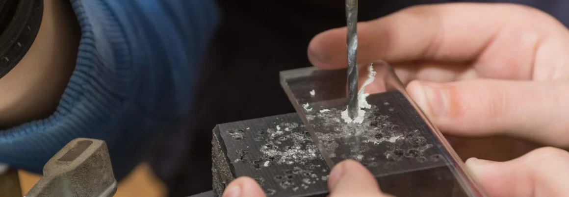 Ein Bohrer bohrt in eine Acrylglasplatte, die von Händen gehalten wird.